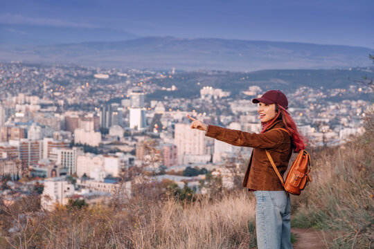 Woman tourist wearing a cap and backpack pointing at the city skyline of Tbilisi, Georgia during autumn