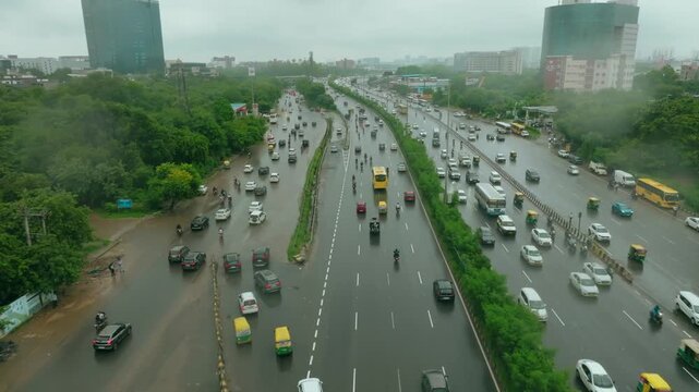 Aerial view of vehicles navigating the wet asphalt of National Highway 48 amidst lush greenery and urban buildings, Gurugram, Haryana, India.