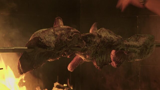 half-roasted suckling pigs on a spit with a wood fire in the background. A hand sprinkles salt and spices.
