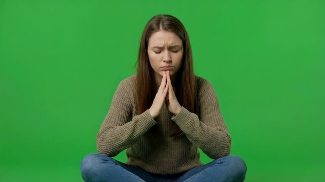 Young woman seated cross legged with hands steeped under chin in deep thought isolated on green screen background emotional concept 4k video footage
