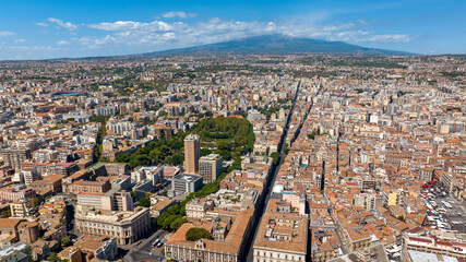 Aerial view of the city center of Catania, Sicily, southern Italy. In the background, on the horizon, Mount Etna looms in silhouette, dominating the panorama. It is a sunny summer day.