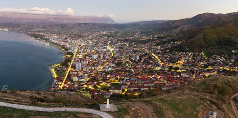 Twilight aerial panorama of Pogradec, Albania, showing illuminated streets, dense neighbourhoods, and the calm shoreline of Lake Ohrid.