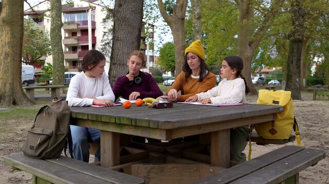 Young female teacher giving a lesson to three students sitting at a wooden picnic table in an outdoor park setting, promoting the concept of alternative education and homeschooling