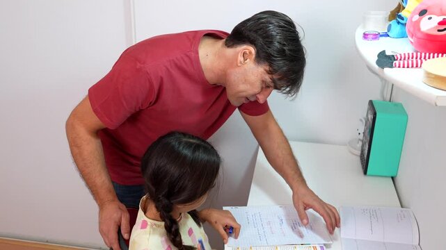 Caring father standing next to his daughter, assisting her with her schoolwork while she sits at her desk, concentrating and writing in her notebook with a pen in a white room
