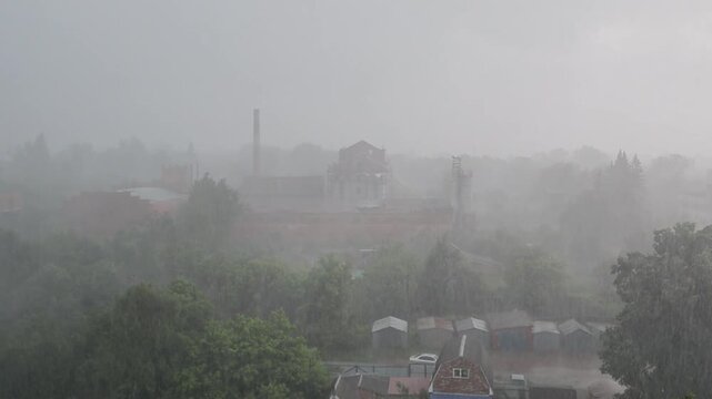 Intense rainfall and thick mist obscure an industrial town, with factory buildings and chimneys barely visible above trees during a gloomy storm.