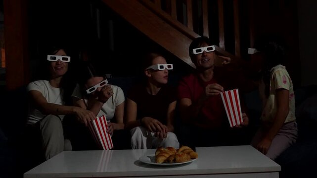 Emotional family with three daughters wearing 3d glasses, eating popcorn, and laughing while watching a comedy movie on the sofa at home, enjoying a fun and entertaining evening together
