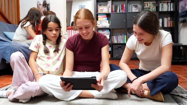 Three happy teenage girls sitting together on the floor in a cozy living room, smiling and interacting while browsing content on a digital tablet, sharing a moment of modern friendship