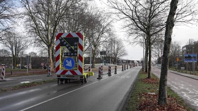 Yellow LED traffic arrow (pijlwagen) trailer on a Dutch road directing vehicles to the right lane during roadworks on a cloudy autumn day with safety cones.
