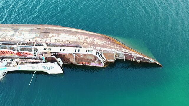 Aerial view of a rusty, capsized ship in the turquoise sea, revealing its weathered hull and lifeboats, a stark contrast against the calm waters, Athina, Kentrikos Tomeas Athinon, Greece.