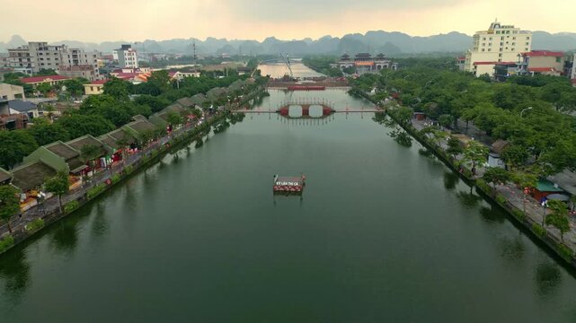 Aerial view of a still river reflecting the sky, flanked by trees and buildings with a bridge in the distance, Hoa Lu, Ninh Binh, Vietnam.