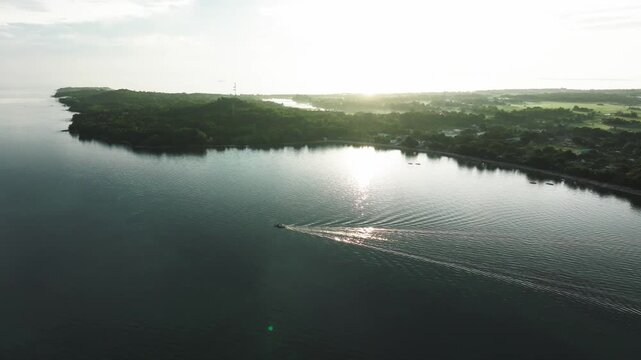 Aerial view of a boat creating a wake in the dark blue water near an island with lush green trees and a bright horizon, Carles, Western Visayas, Philippines.
