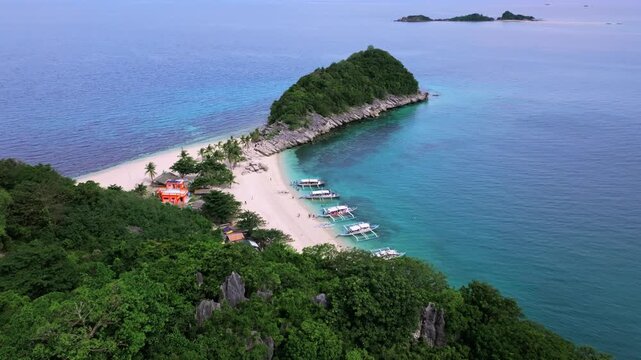 Aerial view of an island with lush green vegetation, white sand beach, docked boats, and clear turquoise water, Carles, Western Visayas, Philippines.