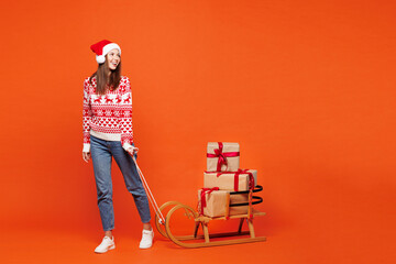 Full body merry fun young woman wear red Christmas sweater Santa hat posing pulling sled with present boxes look aside isolated on plain orange background. Happy New Year celebration holiday concept.
