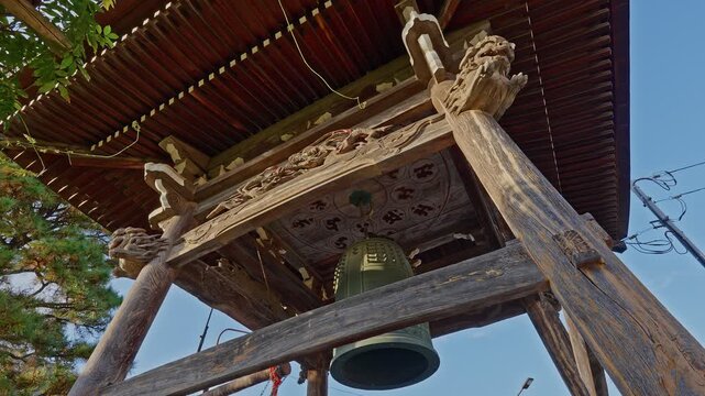 An ancient, intricately carved Japanese Buddhist temple bell (Bonsho) hanging inside a wooden bell tower (Shoro) surrounded by lush forest foliage.