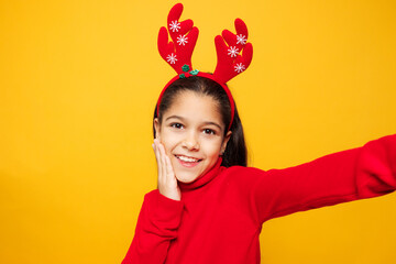 Merry cute little girl wear red clothes fun decorative deer horns on head posing do selfie shot pov on mobile cell phone isolated on plain yellow background. Happy New Year Christmas holiday concept.