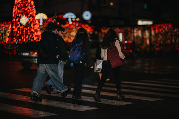 Teenagers walk across a crosswalk at night beneath festive Christmas lights in the city. A group of teens with backpacks move together through a bright, holiday-lit street.