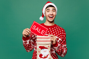 Merry young man wear red cozy Christmas sweater Santa Claus hat posing hold in hand card with sale text in stocking sock isolated on plain green background. Happy New Year celebration holiday concept.