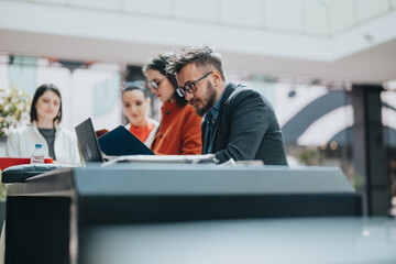 People gather in a bright, contemporary lobby while a man focuses on his laptop. Colleagues discuss documents in the background, creating a collaborative atmosphere.