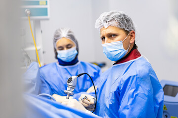 Surgeon Performing Endoscopic Surgery with Assistant. Focused male surgeon using a laparoscopic instrument while his female assistant monitors the process in the background.