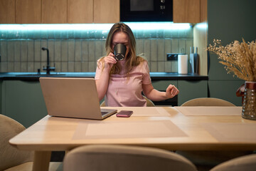 Freelance woman drinking coffee while working on laptop in kitchen