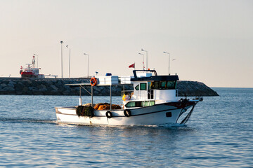 White fishing boat navigating near rocky breakwater—equipped with nets, lifebuoy, and Turkish flag, framed by calm sea, docked vessel, and clear daylight in coastal atmosphere.