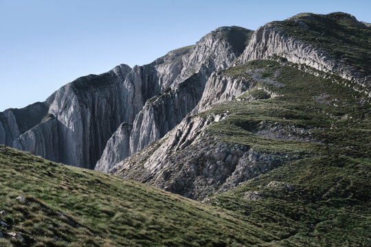Dramatic rocky mountain ridge with steep cliffs and green slopes under clear blue sky, part of the Durmitor range near Prutas, ideal for travel and adventure concepts.