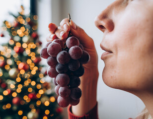 Close-up of woman smelling red grapes with blurred Christmas tree lights