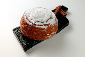 Round, delicious bun with cinnamon and powdered sugar on a black wooden board on a white background
