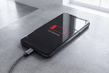 Smartphone charging on a gray concrete surface near window light, close up of mobile device displaying low battery warning with red icon and charging cable connection