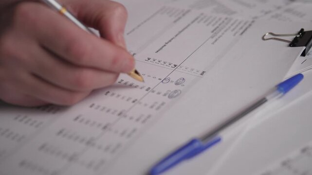 CloseUp Hand Filling Tax Form With Pen, Preparer Marking Boxes And Numbers On Printed Return, Blue Ballpoint Nearby And Binder Clip On Sheet, Careful Calculation For Submission And Refund