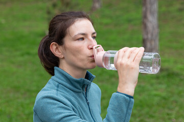 Woman with brown hair wearing a blue fleece jacket is drinking water from a clear bottle while enjoying nature in a green outdoor setting, promoting hydration and wellness © Ekaterina