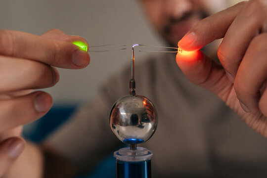 Close-up hands of scientist holding two leds near mini tesla coil, observing electrostatic discharge and electrical sparks illuminating light-emitting diodes during physics experiment, closeup.