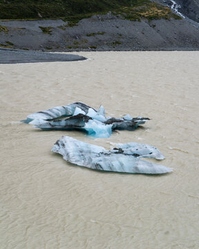 View of luminous blue icebergs adrift in milky waters, reflecting the overcast sky, with rugged mountains in the distance, Hooker Lake, Canterbury Region, New Zealand.