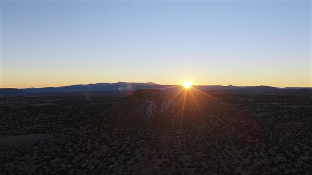 Wide drone shot of a solitary volcanic cinder cone rising from the high desert plains. The low-angle sun creates a dramatic silhouette, highlighting the rugged texture of the New Mexico landscape.