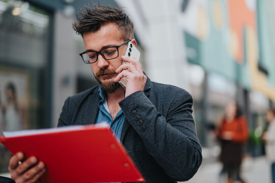 A man wearing glasses and a blazer speaks on a smartphone while holding a red folder in a colorful urban street, among people. He looks focused as he handles work tasks, a sign of busy associates.