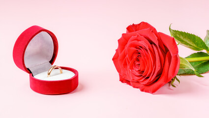 Golden ring tucked in velvet box beside a vibrant red rose on soft pink background