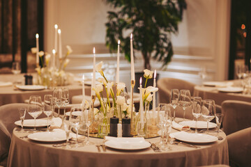 A dining table is prepared for an evening gathering. White flowers sit in the center among candles. Glassware and plates are arranged neatly, creating a formal look for the occasion