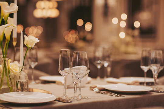 Two glasses stand on a table set for dinner in a restaurant. White flowers and candles add to the atmosphere as light shines softly in the background during the evening
