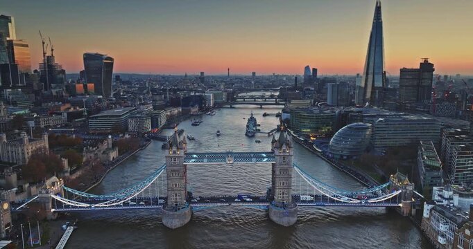 Aerial view Tower Bridge glowing over River Thames at sunset, evening traffic and boats framed by historic landmarks and modern skyscrapers. The Shard and Gherkin night cityscape. Drone flight footage