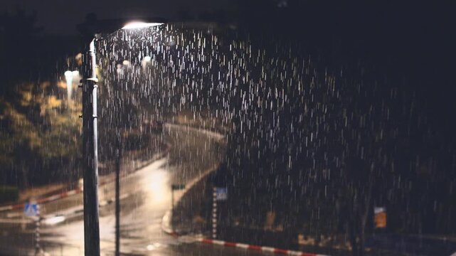 A heavy rain falls at night, illuminated by a lone street lamp. A gloomy and atmospheric scene of a wet road during a thunderstorm. Night rain.