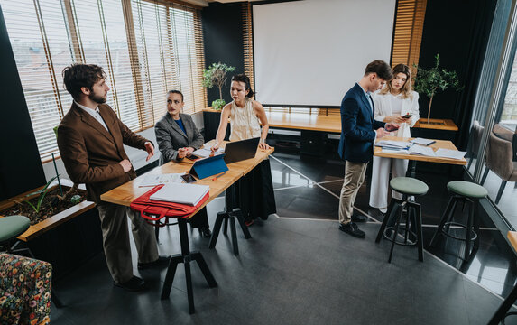 A diverse group of coworkers gather around a tall table with laptops and documents, discussing plans and reviewing materials in a bright, modern office with plants and a projector screen. - Powered by Adobe
