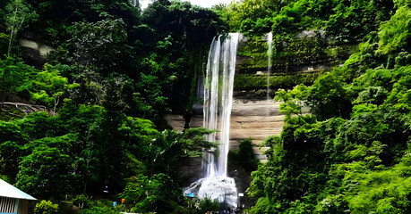 A beautiful natural cascade flows over mossy rocks in a lush green forest park, creating a stunning tropical landscape where water streams through the trees
