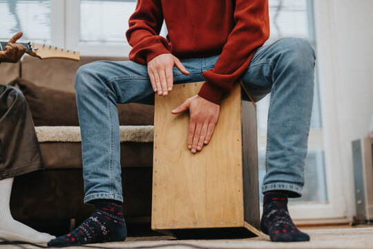 A person sits on a cajon drum in a living room, playing with hands. A casual indoor music session with a guitar nearby and relaxed, cozy atmosphere.
