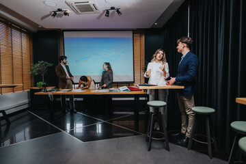 A group of business coworkers collaborates in a contemporary conference room around a long table. A projector shows charts while colleagues stand and talk, sharing ideas and planning together.