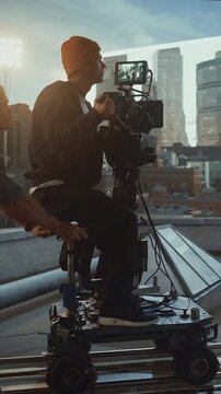 Vertical Screen: Camera Operator in Beanie Rides a Rolling Dolly on Rooftop Set, Steering Professional Digital Cinema Rig and Recording Smooth Tracking Shots for a Narrative Movie Scene