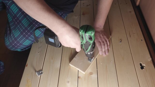 Overhead close-up of hands using a cordless drill to fasten a metal bracket to a wooden block on a plank table. Indoor DIY woodworking and repair scene