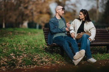 Fototapeta premium A man and woman sit on a park bench, talking and smiling. They share a casual moment outdoors in a calming autumn park setting.