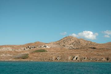 Clear coastal view of Delos island showing scattered ancient ruins on a rocky hillside, bright blue sky, and deep turquoise water below.
