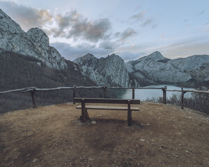 View of a solitary wooden bench poised on a precipice overlooking a serene lake embraced by rugged mountains under a pastel sky, Riano, Castilla y León, Spain.