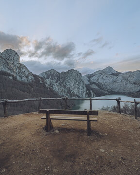 View of a wooden bench sits overlooking a serene lake surrounded by rugged, snow-dusted mountains under a pastel sky, Riano, Castilla y Le&oacute;n, Spain.